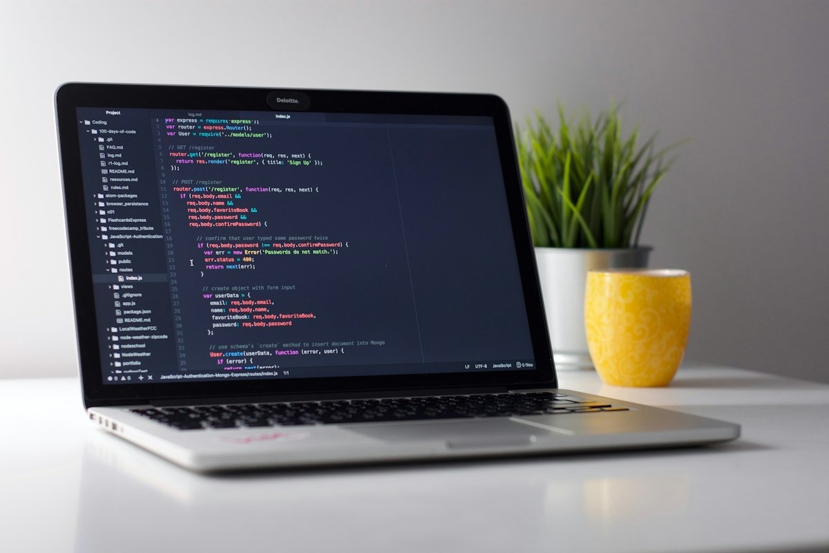 A laptop showing website code beside a small plant and a yellow mug